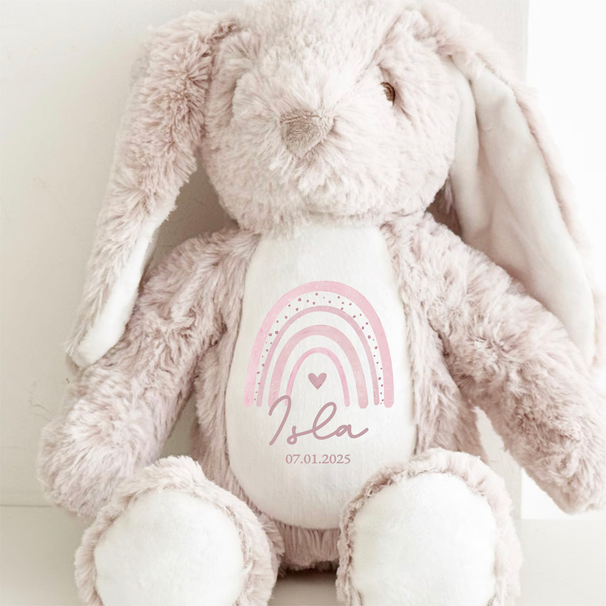 Beige soft bunny toy with personalised name and pink rainbow print on the front. A timeless keepsake by Ayla & Lara, perfect for baby gifts.