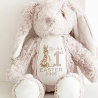 A soft toy Beige Bunny with long ears sitting up with a Peter Rabbit Baby's First Easter 2025 printed on the front.